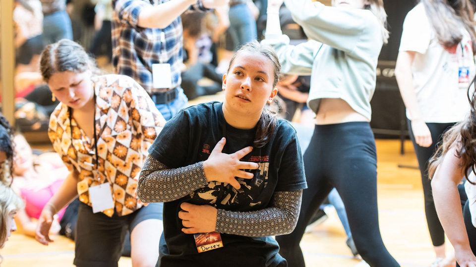 A girl aged about twelve years old in a black tee shirt is dramatically wrapping her hands around her torso. Other young people are in the background acting out a scene.
