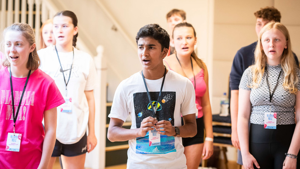 A group of young people in tee shirts stood with their mouths open as they learn vocal techniques.