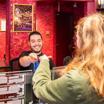 A male Box Office Supervisor in a black shirt handing a ticket to a female customer with blonde hair and a green blouse