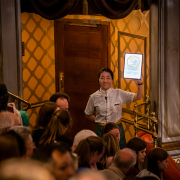 A female member of staff stood inside the auditorium with audience members in their seats. She is wearing a white blouse and is holding up an ipad displaying a no photography sign