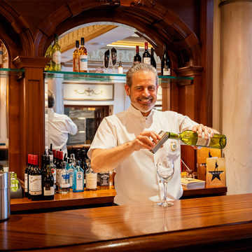 A male bar supervisor in a white shirt stood behind a bar pouring out a measure of wine