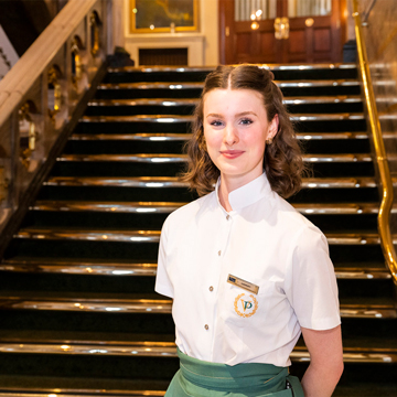 Female member of staff in a white blouse and green apron, stood at the foot of stairs in the theatre foyer, smiling
